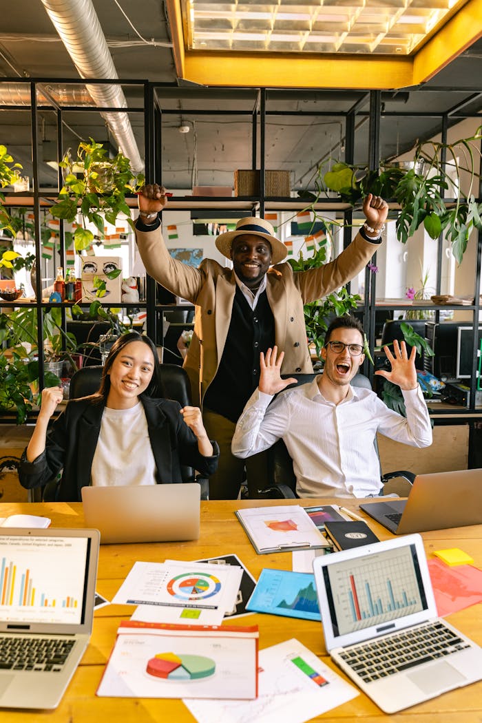 Happy diverse team celebrating success in a modern office with laptops and charts.